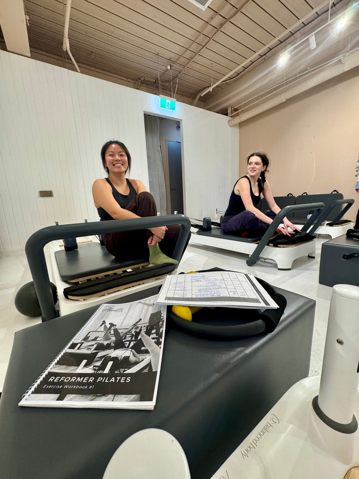 Two women using Pilates reformers in a studio with a Pilates reformer manual on a table.